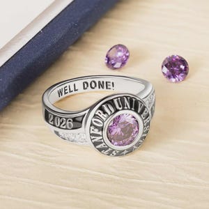 May include: A silver ring with a large, round, purple gemstone at the center. The ring is engraved with "WELL DONE!" and "2026". Two smaller purple gemstones are scattered nearby. The ring is likely a graduation or class ring.