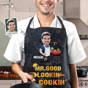 Könnte beinhalten: Schwarze Schürze mit einer Cartoon-Karikatur eines grillenden Mannes, dem Namen "Michael" und dem Text "Mr. Good Lookin' Cookin'". Die Schürze hat eine weiße Latzhose und ist mit Illustrationen zum Thema Essen verziert. Ein Foto der Person befindet sich in der oberen linken Ecke.