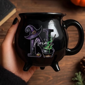 May include: Black cauldron-shaped mug with a witch hat and the name "Nora" in a stylized font. The design includes a cauldron with green potion, a witch hat, and a pumpkin. The mug has a handle and three small feet.