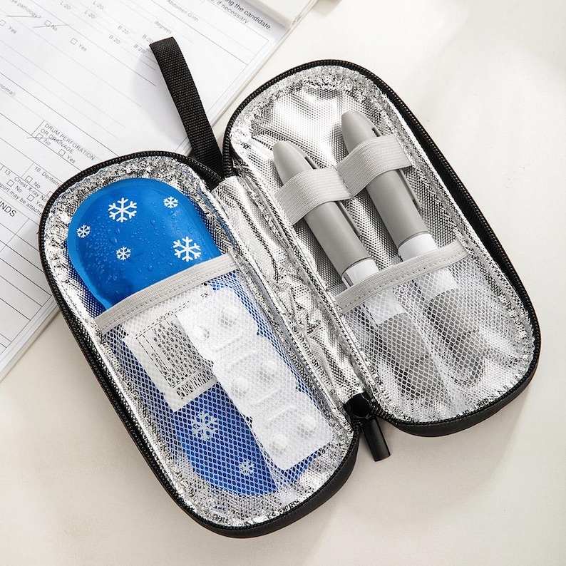 May include: Open black insulated medical travel case with a zipper. Inside, there's a blue ice pack with white snowflakes, a blister pack of pills, and two gray medical devices secured with elastic straps. The interior is silver.