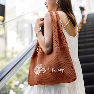 Puede incluir: Un bolso tote de ganchillo de color óxido con un bordado floral y el nombre "Tracey" en cursiva. El bolso tiene un diseño de tejido abierto grande y se lleva al hombro. El bolso está hecho de un material suave y texturizado.