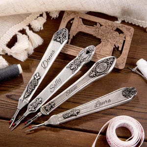 May include: Four silver seam rippers with ornate floral designs and personalized names: Donna, Sally, Elaine, and Laura. A wooden sewing machine-shaped decoration and a measuring tape are also visible.