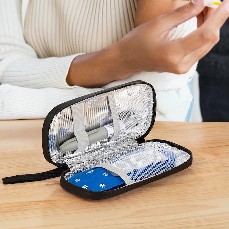 May include: Black insulated medical travel case, open to reveal a gray medical pen and a blue ice pack with white snowflake designs. The case has a silver insulated lining and a black zipper.