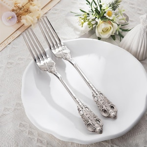 May include: Two silver forks with ornate handles rest on a white plate. One fork is engraved with "Mr." and the other with "Mrs." and the date "10.16.23". The forks are part of a wedding or anniversary set.