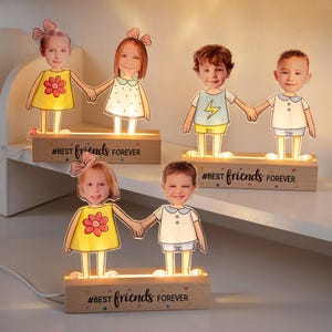 Puede incluir: Luces de noche de madera personalizadas con caras de niños tomados de la mano. Cada luz tiene un brillo cálido y el texto "#BEST friends FOREVER" en la base. Los niños visten trajes coloridos.