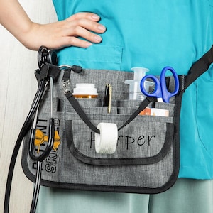 May include: A grey medical utility belt with multiple pockets, holding a stethoscope, scissors, pen, tape, and other medical supplies. The belt has the name "Harper" printed on it.