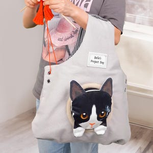 May include: A light gray tote bag with a black and white cat design. The bag has a circular opening with a plush cat face and the text "Bella's Project Bag". The bag is being used for a knitting project.
