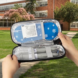 May include: Black insulated medical case, open to reveal a blue ice pack with white snowflakes and two gray medical devices secured with gray straps. The case is held open against a blurred outdoor background.