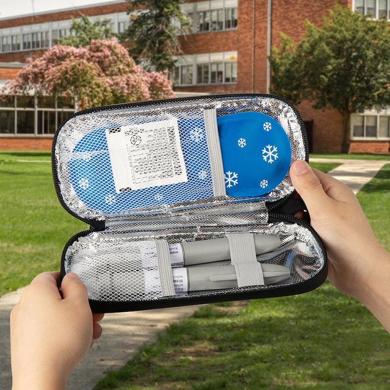 May include: Black insulated medical case, open to reveal a blue ice pack with white snowflakes and two gray medical devices secured with gray straps. The case is held open against a blurred outdoor background.
