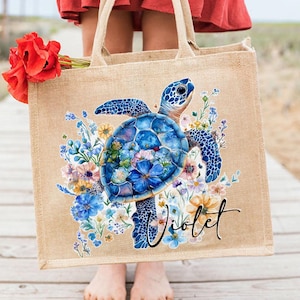 Puede incluir: Bolso tote de yute beige con un diseño de tortuga marina azul rodeada de flores coloridas. La bolsa tiene el nombre "Violet" en escritura negra y asas resistentes. Un ramo de flores rojas descansa en el lateral.