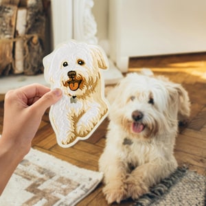 Puede incluir: Un parche bordado con la forma de un perro blanco se sostiene frente a un perro vivo de la misma raza. El parche tiene un diseño detallado con acentos marrones y beige, que representa la cara y el cuerpo del perro. El perro vivo está acostado con la lengua fuera.