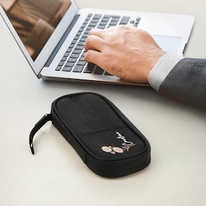 May include: A black rectangular pencil case with a zipper closure and a small handle. The case has a floral design and the name "Caroline" embroidered on the front. A person is typing on a laptop in the background.