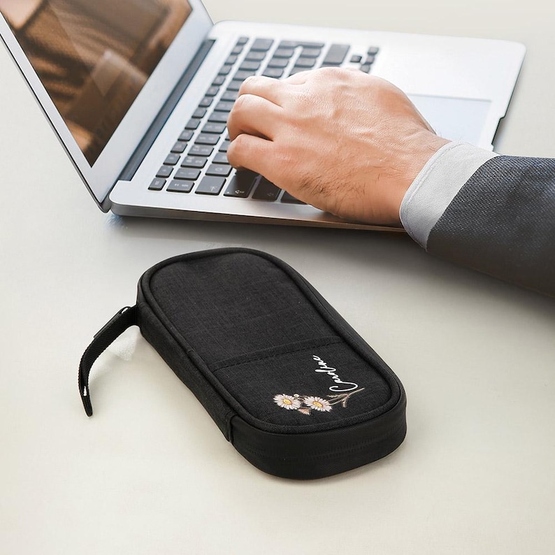 May include: A black rectangular pencil case with a zipper closure and a small handle. The case has a floral design and the name "Caroline" embroidered on the front. A person is typing on a laptop in the background.