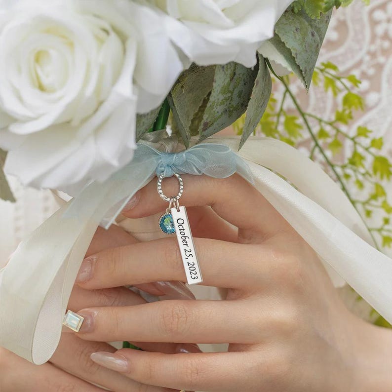May include: A bouquet of white roses with a light blue ribbon. A silver charm with a small blue and green globe and a rectangular tag engraved with "October 25, 2023" hangs from the ribbon.