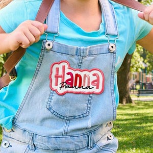 May include: Light blue denim overalls with a patch that reads "Hanna" in a cursive font with a red and white border. The overalls have adjustable straps and metal hardware. A light blue t-shirt is worn underneath.