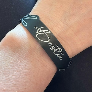 May include: A black wristband with the word "Bestie" in white script. The wristband has a decorative floral design on the edges. The wristband is worn on a wrist.