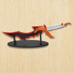 May include: A decorative dagger with a black and red blade, orange hilt, and silver handle, displayed on a black oval stand. The blade has a unique, stylized shape. The dagger is set against a light brown wooden background.