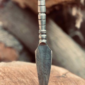 May include: A silver-colored, hand-forged, spear-shaped knife with a textured handle. The knife is sharp and pointed, and the handle is made of metal.