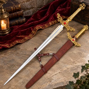 May include: A silver-toned metal sword with a gold-colored hilt featuring intricate filigree, a central red gemstone, and a crown-shaped pommel. The sword is accompanied by a brown leather scabbard with metal hardware and a matching leather harness.