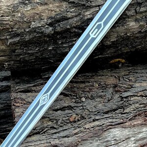 May include: A silver sword blade with black and white decorative lines and geometric designs. The blade is angled across a textured, brown wooden surface. The sword's design features a series of parallel lines and stylized symbols.
