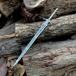May include: A silver-colored sword with a detailed hilt and a long, pointed blade. The sword features intricate designs along the blade and hilt. The sword is resting on a pile of rough-hewn logs, creating a rustic aesthetic.