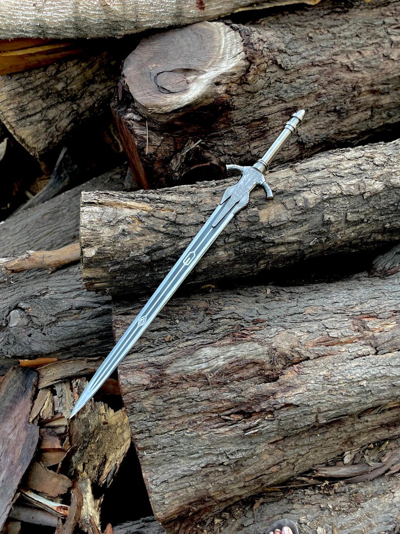 May include: A silver-colored sword with a detailed hilt and a long, pointed blade. The sword features intricate designs along the blade and hilt. The sword is resting on a pile of rough-hewn logs, creating a rustic aesthetic.
