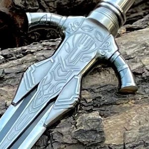 May include: Close-up of a silver sword with intricate designs on the hilt and blade. The hilt features a stylized panther head, and the blade has a central groove. The sword is set against a textured, brown background.