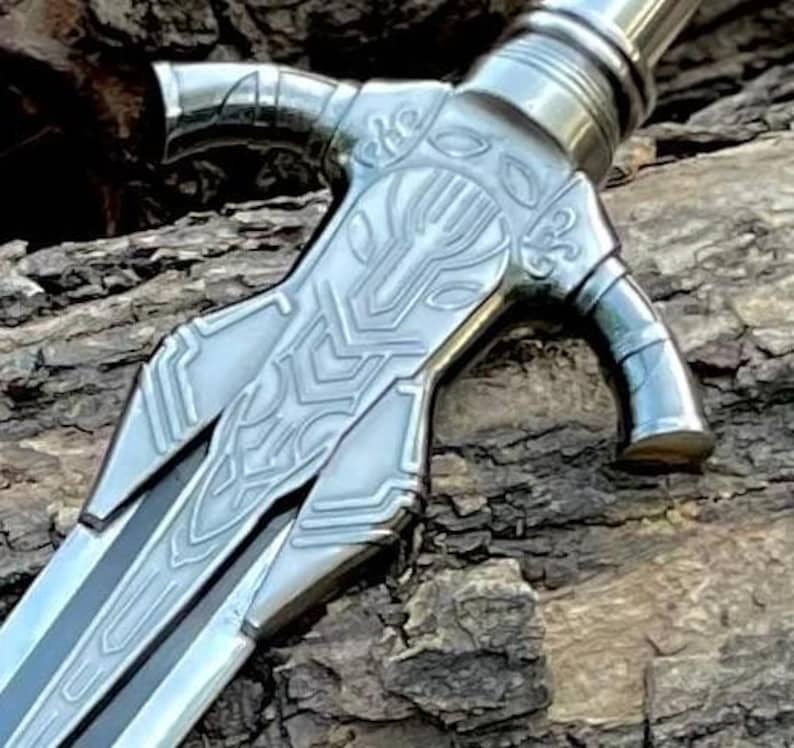 May include: Close-up of a silver sword with intricate designs on the hilt and blade. The hilt features a stylized panther head, and the blade has a central groove. The sword is set against a textured, brown background.