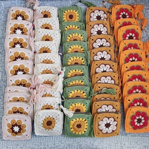 May include: A collection of handmade crochet coin purses in various colors, including cream, pink, green, yellow, and orange. Each square purse features a sunflower design and a tassel. The purses are arranged in neat rows.