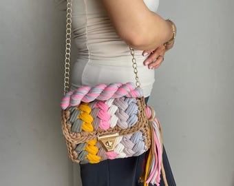 May include: A colorful crocheted crossbody bag with a gold chain strap and a tassel. The bag is made of different colored yarn, including pink, blue, green, and yellow.