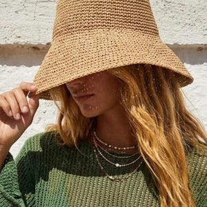 Könnte beinhalten: Ein beigefarbener gewebter Eimerhut mit breiter Krempe. Der Hut wird mit einem grünen Strickpullover und mehrschichtigen goldenen Halsketten getragen. Der Hut besteht aus einer Naturfaser und hat ein strukturiertes Aussehen.