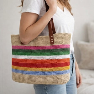 May include: A woven tote bag with a beige body and horizontal stripes in rainbow colors. The bag has brown leather handles. The bag is held by a person wearing a white t-shirt and blue jeans.