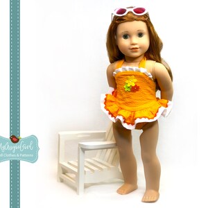 Puede incluir: Una mu&ntilde;eca con un traje de ba&ntilde;o naranja con ribete de volantes blancos y un adorno floral. La mu&ntilde;eca tiene el pelo largo y rojo y lleva gafas de sol blancas. Detr&aacute;s de la mu&ntilde;eca hay una silla de madera blanca. El texto "MyAngieGirl Doll Clothes & Patterns" est&aacute; a la izquierda.