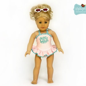 Puede incluir: Una mu&ntilde;eca con un traje de ba&ntilde;o a rayas rosas y blancas con una falda con volantes y ribete turquesa. El traje de ba&ntilde;o tiene un monograma y se combina con gafas de sol blancas. La mu&ntilde;eca tiene el pelo rubio.