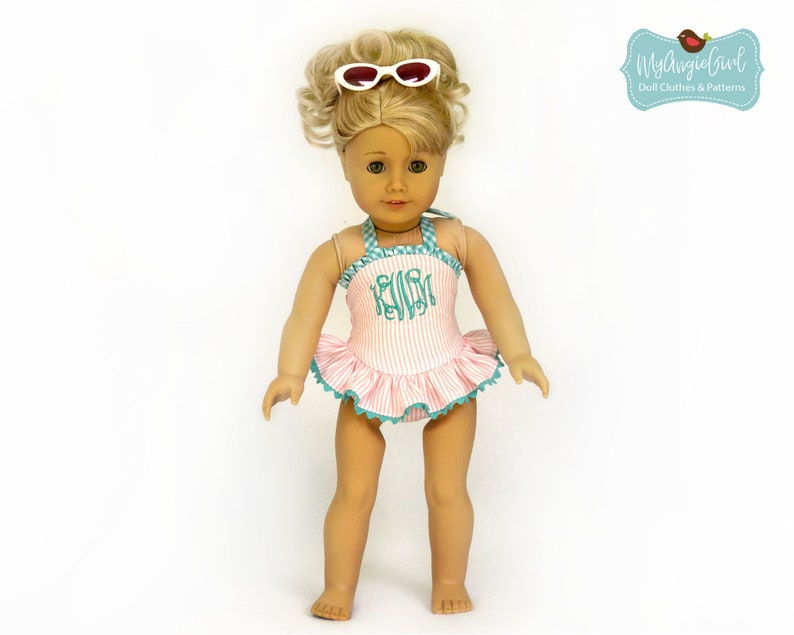 Puede incluir: Una mu&ntilde;eca con un traje de ba&ntilde;o a rayas rosas y blancas con una falda con volantes y ribete turquesa. El traje de ba&ntilde;o tiene un monograma y se combina con gafas de sol blancas. La mu&ntilde;eca tiene el pelo rubio.