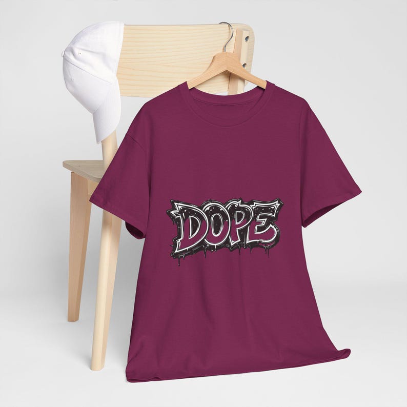 May include: A maroon t-shirt with the word "DOPE" in a graffiti-style design. The word is outlined in white with black shading and dripping accents. A white baseball cap rests on a wooden chair.
