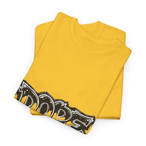 May include: Two folded yellow t-shirts. The bottom of the top shirt has the word "DOPE" in a black and white graffiti-style font. The shirts are made of a soft material and are folded neatly.