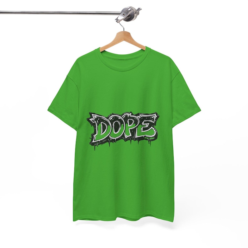 May include: A bright green t-shirt with the word "DOPE" in a graffiti-style design. The word is outlined in white and black, with green fill and dripping paint effect. The shirt is hanging on a wooden hanger.