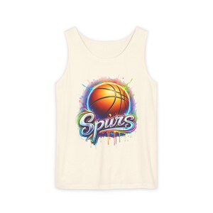 May include: Cream-colored tank top featuring a basketball graphic with the word "Spurs" in a colorful, stylized font. The basketball is orange with black lines, set against a vibrant, splashy background of blues, greens, and purples.