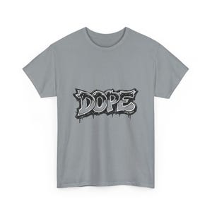 May include: A light gray t-shirt featuring the word "DOPE" in a graffiti-style design. The word is rendered in black and white with dripping paint effects, creating a bold and urban aesthetic. The shirt is a classic crew neck style.