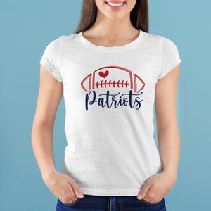 May include: White t-shirt featuring a red and blue football design with a heart and the word "Patriots" in a script font. The shirt is a classic crew neck style.