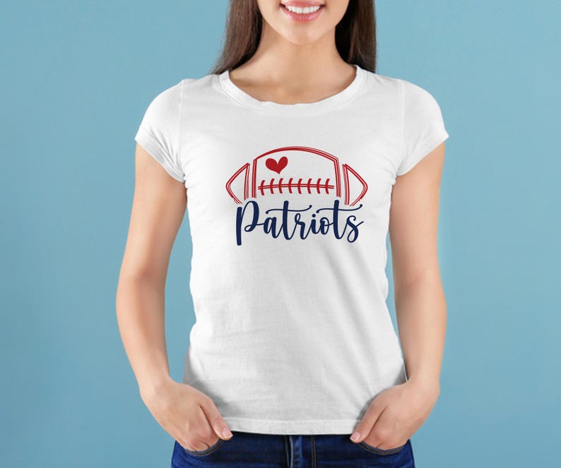 May include: White t-shirt featuring a red and blue football design with a heart and the word "Patriots" in a script font. The shirt is a classic crew neck style.
