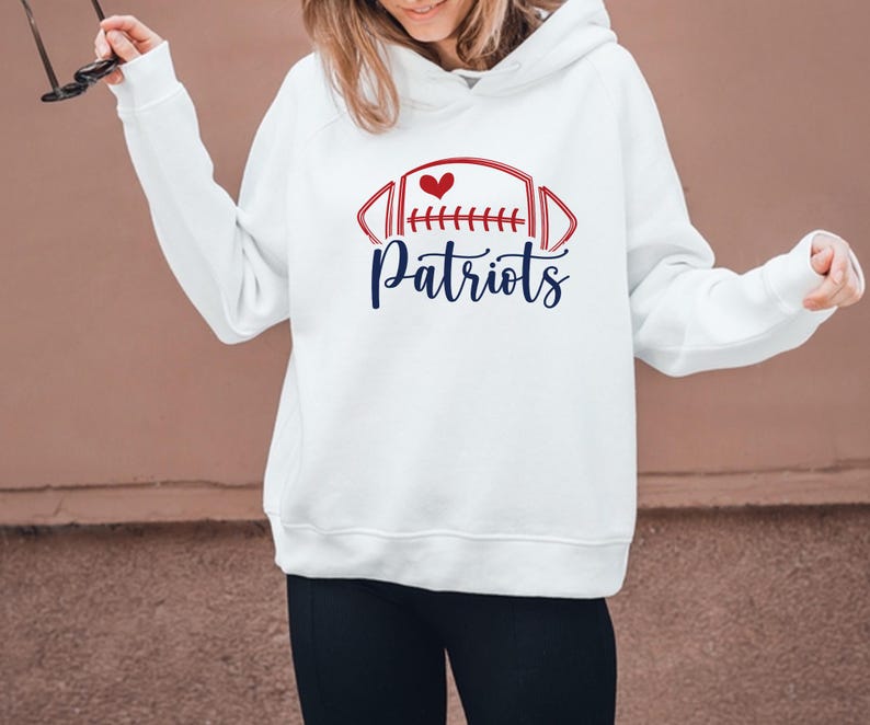 May include: White hoodie with a red and blue football graphic and the word "Patriots" in a script font. The graphic includes a red heart. The hoodie has long sleeves and a hood.