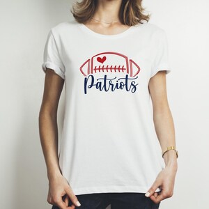 May include: White t-shirt featuring a red and blue football design with a heart and the word "Patriots" in a script font. The shirt is a classic crew neck style.