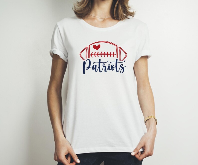 May include: White t-shirt featuring a red and blue football design with a heart and the word "Patriots" in a script font. The shirt is a classic crew neck style.