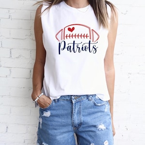 May include: White sleeveless shirt with a red and blue football graphic and the word "Patriots" in a script font. The graphic includes a heart. The shirt is paired with denim shorts.