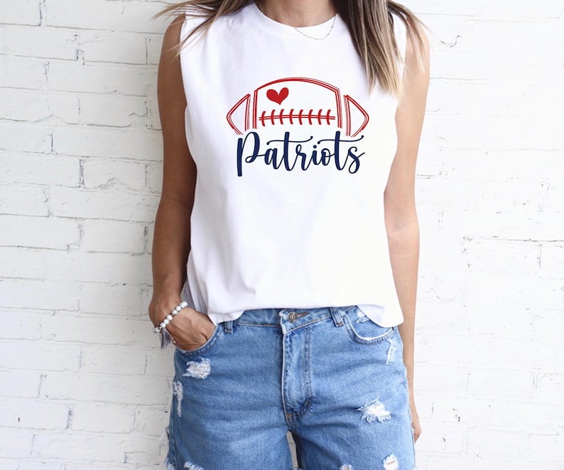 May include: White sleeveless shirt with a red and blue football graphic and the word "Patriots" in a script font. The graphic includes a heart. The shirt is paired with denim shorts.