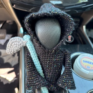 Puede incluir: Muñeca de la Parca hecha a mano con ganchillo, con una capa con capucha negra y una guadaña gris. La muñeca tiene motas de colores en el hilo y está colocada en la palanca de cambios de un coche.