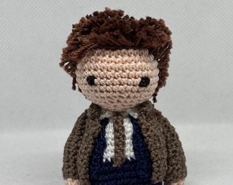 Puede incluir: Una muñeca de crochet con pelo marrón, una chaqueta marrón, una camisa blanca y pantalones azules.