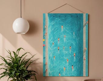 Op de afbeelding: Een rechthoekige wandkunstprint met een zwembad vanuit vogelperspectief. Het turquoise water is gevuld met zwemmers in kleurrijke zwemkleding. Het kunstwerk hangt aan een beige muur, met een witte globe hanglamp en een groene plant op de voorgrond.